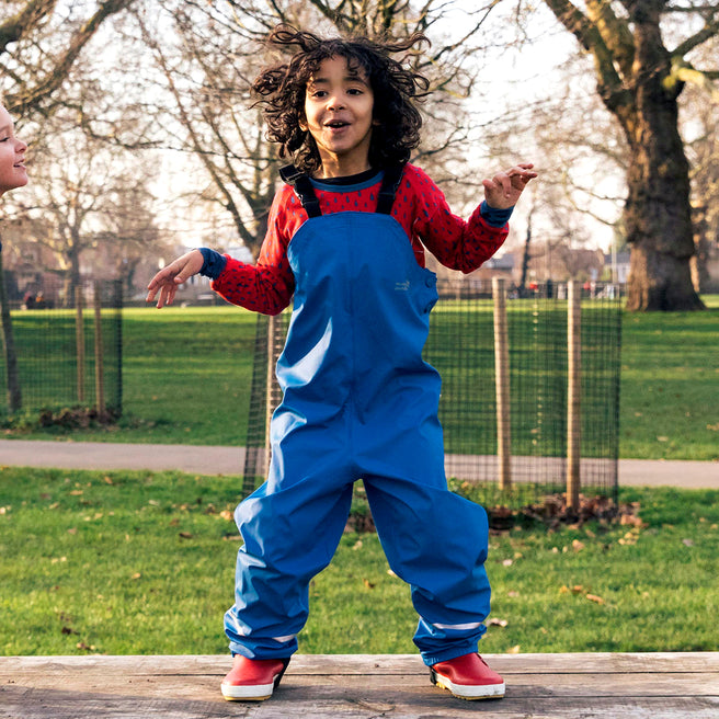 A child with curly hair leaps on a wooden platform in a park, wearing blue waterproof dungarees and red boots.
